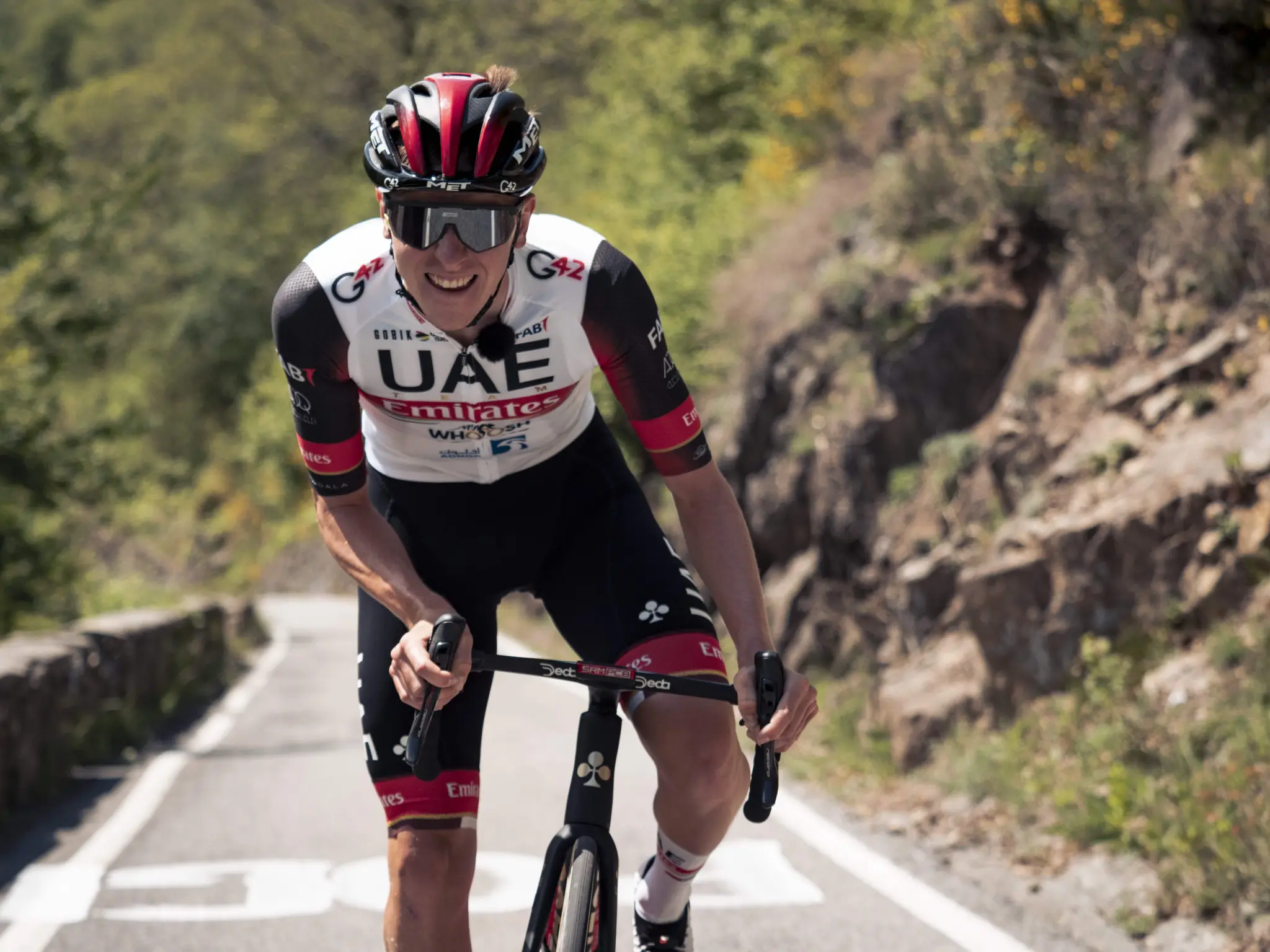 A rider in a UAE Emirates jersey climbs a mountain road, wearing a MET helmet